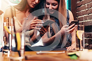 Two cheerful girlfriends using phone looking at screen smiling while having breakfast at coffee house