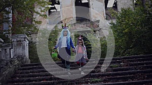 Two cheerful little girls, in colorful costume of unicorn, blue hair and green halloween bucket, walking on abandoned stairs.