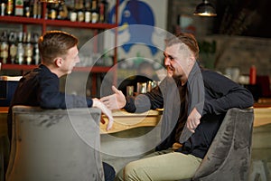 Two cheerful men of friends sit in a bar at the bar and talk about something laughing.