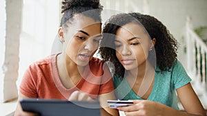 Two cheerful mixed race curly girlfriends shopping online with tablet computer and credit card at home