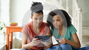 Two cheerful mixed race curly girlfriends shopping online with tablet computer and credit card at home