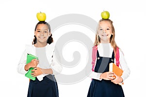 two cheerful multicultural schoolgirls with apples on heads holding books isolated on white.