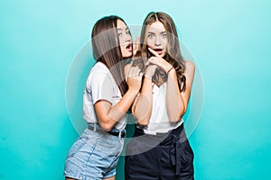 Two cheerful multiethnic young girls dressed in summer clothes whispering secrets over blue background