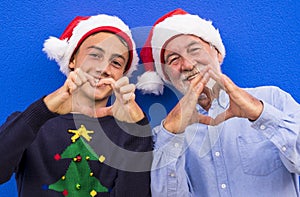 Two cheerful Santa Clauses, a grandfather with his teenage grandson, smile making a heart with their hands. Concept of family,
