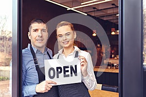 Two cheerful small business owners at entrance of newly opened pub restaurant holding open sign board