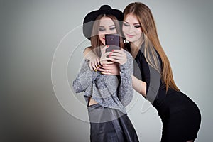 Two cheerful smiling girl girlfriends photographed on the phone, do selfie phone in the studio on a gray background