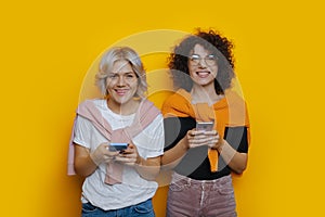 Two cheerful smiling students with curly hair using their phones are posing happily on a yellow background