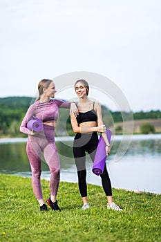 Two cheerful sportswomen in tracksuits talking and laughing while standing with yoga mat by lake in morning