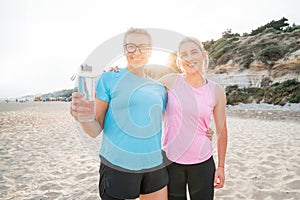 Two cheerful sporty active middle aged women smiling at camera together standing on the beach after workout training on