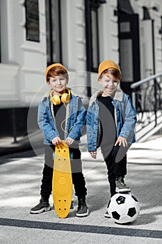 Two cheerful twin boys posing with happy faces in the street. Skateboard or pennyboard and soccer ball.