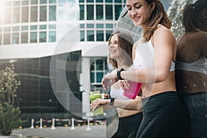 Two cheerful women runners stand leaning against trailer, rest after training, drink water, looking on pulsometer.