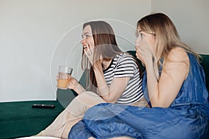 Two cheerful women sitting on green comfortable sofa in living room, together watching humorous TV show and having fun