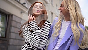 Two cheerful young beautiful woman friends hugging, laughing and greeting waving hands looking at camera standing in