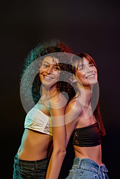 Two cheerful young brunette female models with professional art makeup smiling, posing together in neon light isolated