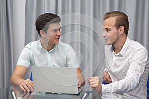 Two cheerful young businessmen working and using laptop on business meeting together