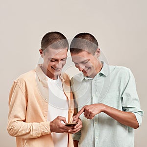 Two cheerful young twin brothers in casual wear using smartphone app while standing together isolated over beige