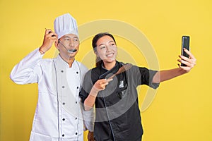 two chefs holding cooking utensils while selfie using mobile phones