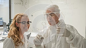 Two chemist colleagues write formulas on glass. Caucasian elderly man and young woman brainstorming.