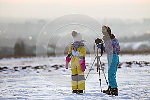 Two children boy and girl having fun outside in winter playing with photo camera on a tripod on snow covered field