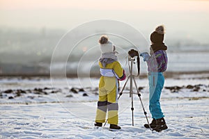 Two children boy and girl having fun outside in winter playing with photo camera on a tripod on snow covered field