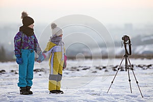 Two children boy and girl having fun outside in winter playing with photo camera on a tripod on snow covered field