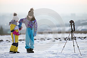 Two children boy and girl having fun outside in winter playing with photo camera on a tripod on snow covered field