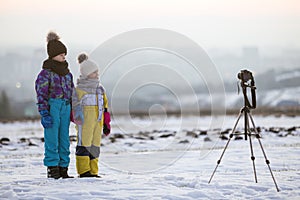 Two children boy and girl having fun outside in winter playing with photo camera on a tripod on snow covered field