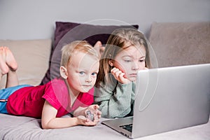 Two children a boy and a girl lie on a sofa in home clothes and watch videos on a laptop