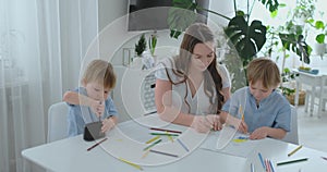 Two children of boys draw with his mother sitting in the kitchen. Happy family at home. Brothers draw at the table. Real