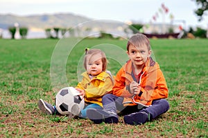 Two children in bright jackets, brother and sister, sit on the football field with soccer ball after game