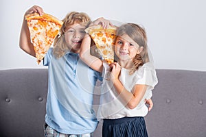 Two children eating pizza and smiling indoors. Happy smiling kids holding pizza slice near face.