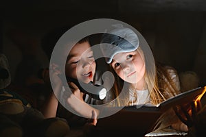 Two children with flashlight read a book under a blanket as a tent