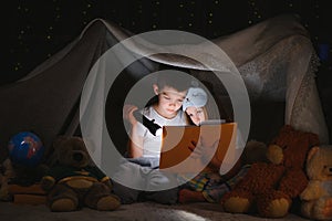 Two children with flashlight read a book under a blanket as a tent