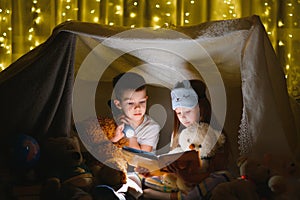 Two children with flashlight read a book under a blanket as a tent.