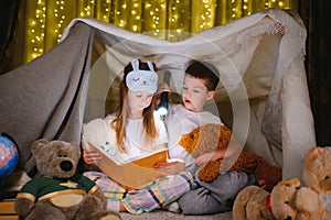 Two children with flashlight read a book under a blanket as a tent.