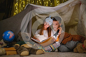 Two children with flashlight read a book under a blanket as a tent.