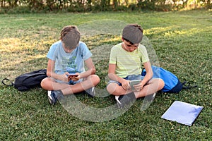 Two children look at their cell phones while sitting with their backpacks in a park. View from above