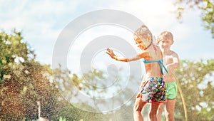 Two children play with sprinkling water in summer garden