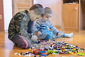 Two children playing with lots of colorful plastic blocks constructor sitting on a floor indoor. Two little brothers play