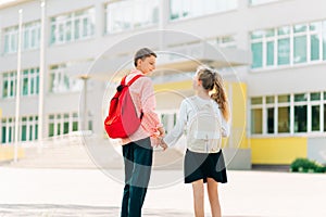 Two children are ready for primary school, a boy and a girl go hand in hand to lessons with backpacks, near the school building