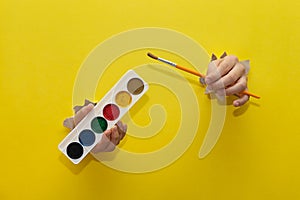 Two children\'s hands, stretched through torn sheet of yellow paper, hold brushes and paints for drawing