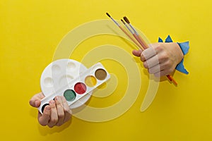 Two children\'s hands, stretched through torn sheet of yellow paper, hold brushes and paints for drawing