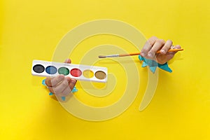 Two children\'s hands, stretched through torn sheet of yellow paper, hold brushes and paints for drawing