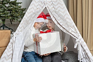 Two children in Santa hats open a gift for Christmas and New Year sitting in a tent-wigwam in the children& x27;s room