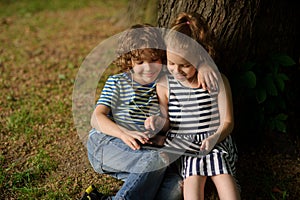 Two children sit under more tree with interest looking at the tablet screen.