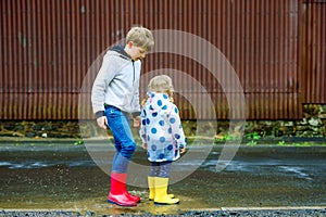Two children, toddler girl and kid boy wearing red and yellow rain boots, walking during sleet. Happy siblings, brother