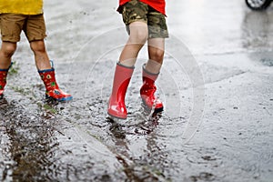 Two children wearing red rain boots jumping into a puddle.