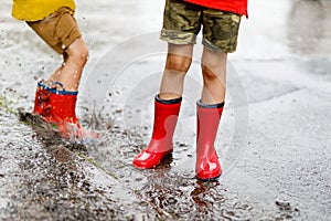 Two children wearing red rain boots jumping into a puddle.