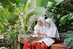two children wearing veil in school uniforms using a cellphone and a book while studying together