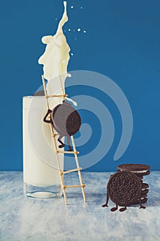 Two chocolate cookies climb stairs into glass of milk on blue paper background. Creative concept. Still life